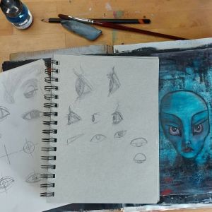 Three sketchbooks on a wooden table. Two sketchbooks are open at pages with practise sketches of eyes in several styles and angles. The third book is open showing a page with grungey turquoise and black paint and a stylised alien-like face with piercing eyes. A pencil, brush, putty rubber and bottle of ink sit on the table above the sketchbooks.